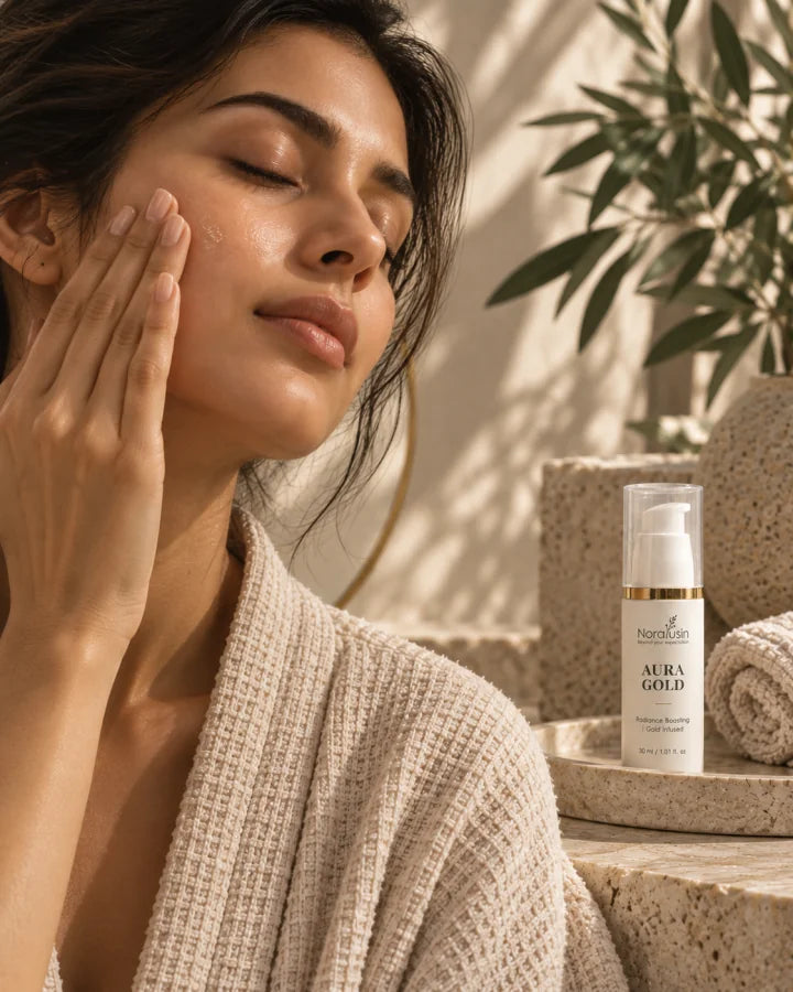 A woman with closed eyes smoothing skincare along her cheek, with a bottle of Aura Gold on a stone tray beside a rolled towel and an olive branch